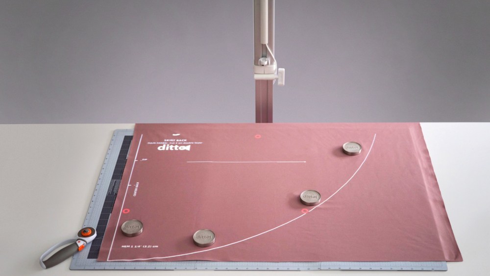 A Ditto digital projector and pattern-drafting workstation featuring a mauve piece of fabric draped across a pattern-making mat with tools scattered atop neatly.